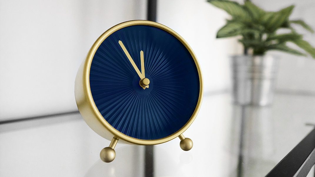Small golden tabletop clock with blue analog face on glass shelf. Popular round clock with hour and minute hands on glossy surface in office. Decorative mantel clock on rack at home. Quartz movement